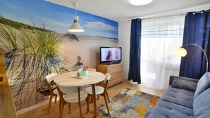 A living room with a beach mural, a dining area with a round table and chairs, a sofa, a TV, and a lamp. - Cozy Holiday Apartment Close to the Beach (Kołobrzeg)