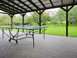 An outdoor ping pong table under a covered pavilion with a brick floor, surrounded by a grassy area with trees and a picnic table.