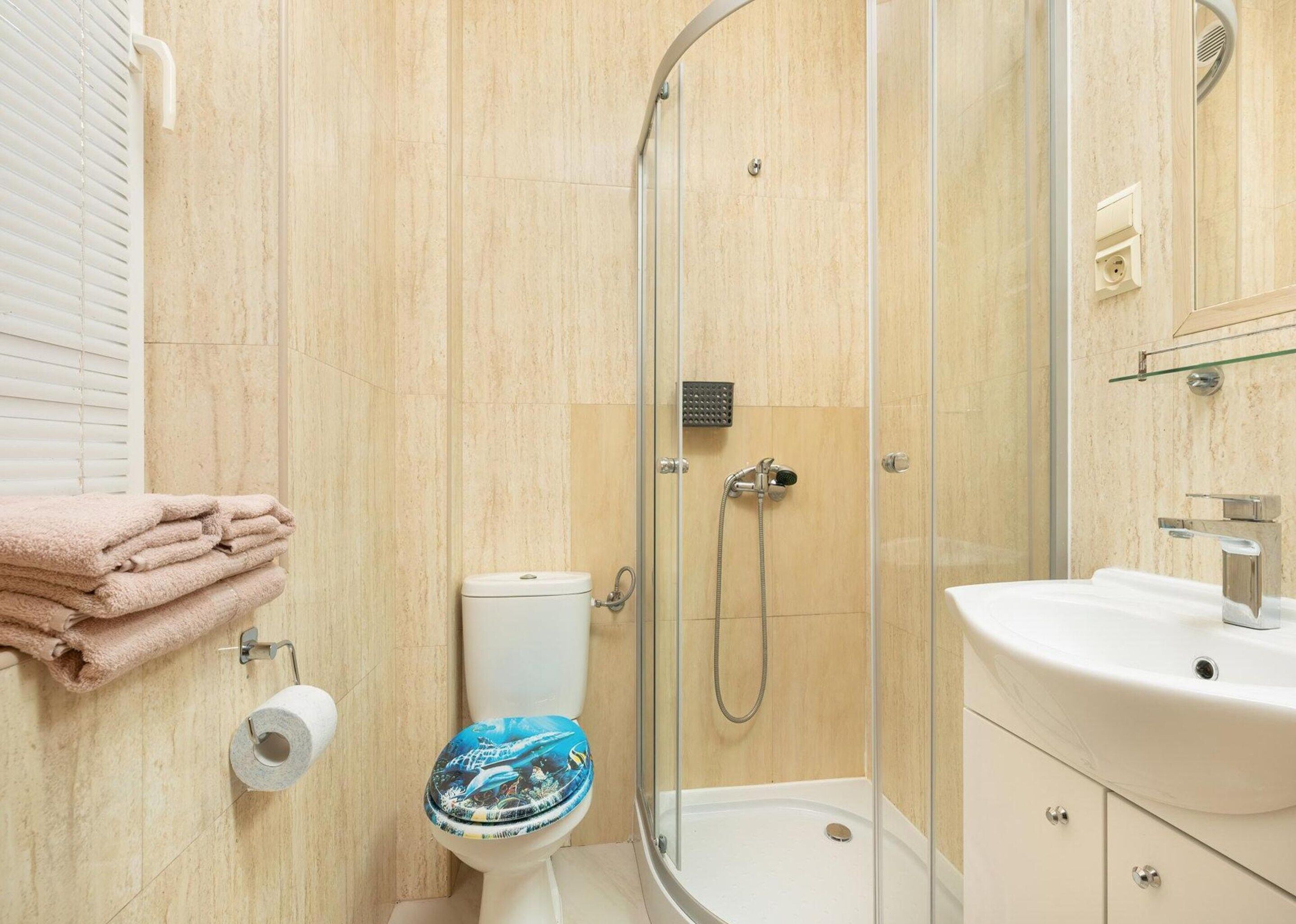 A modern bathroom with a glass-enclosed shower, a toilet with a blue lid, and a sink with a mirror above it.