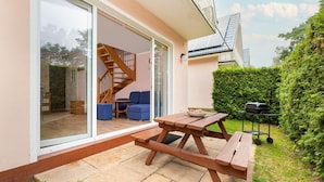 Une terrasse avec une table et des bancs en bois, un barbecue et un escalier menant à un espace intérieur où se trouve un canapé bleu.
