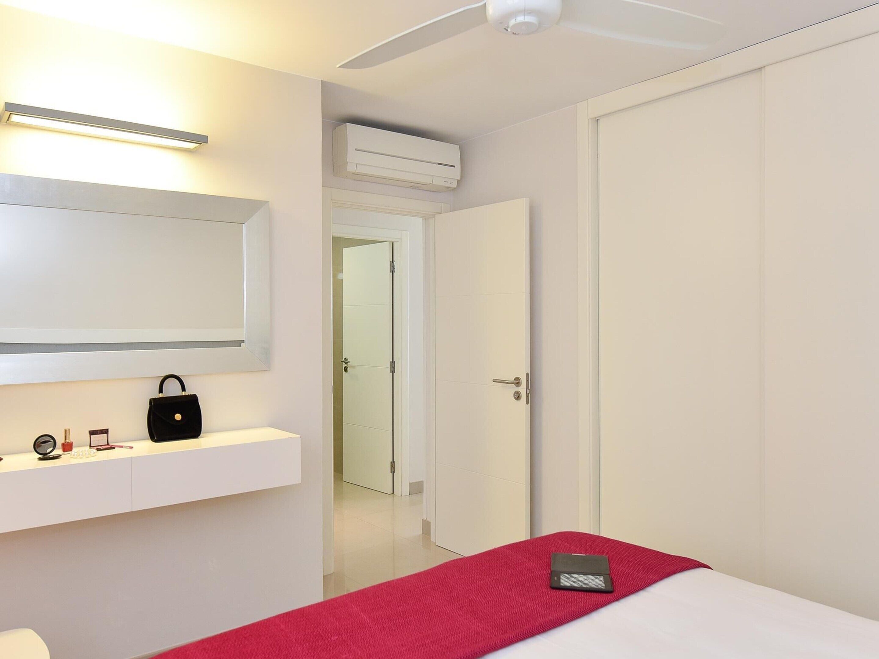 Una habitación de hotel con una cama, una mesita de noche con una manta roja, una pared blanca con un espejo y un equipo de aire acondicionado.