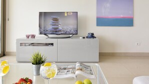 Ein modernes Wohnzimmer mit einem Flachbildfernseher auf einem weißen Schrank, einer Zeitschrift auf einem Glastisch und einer Topfpflanze.