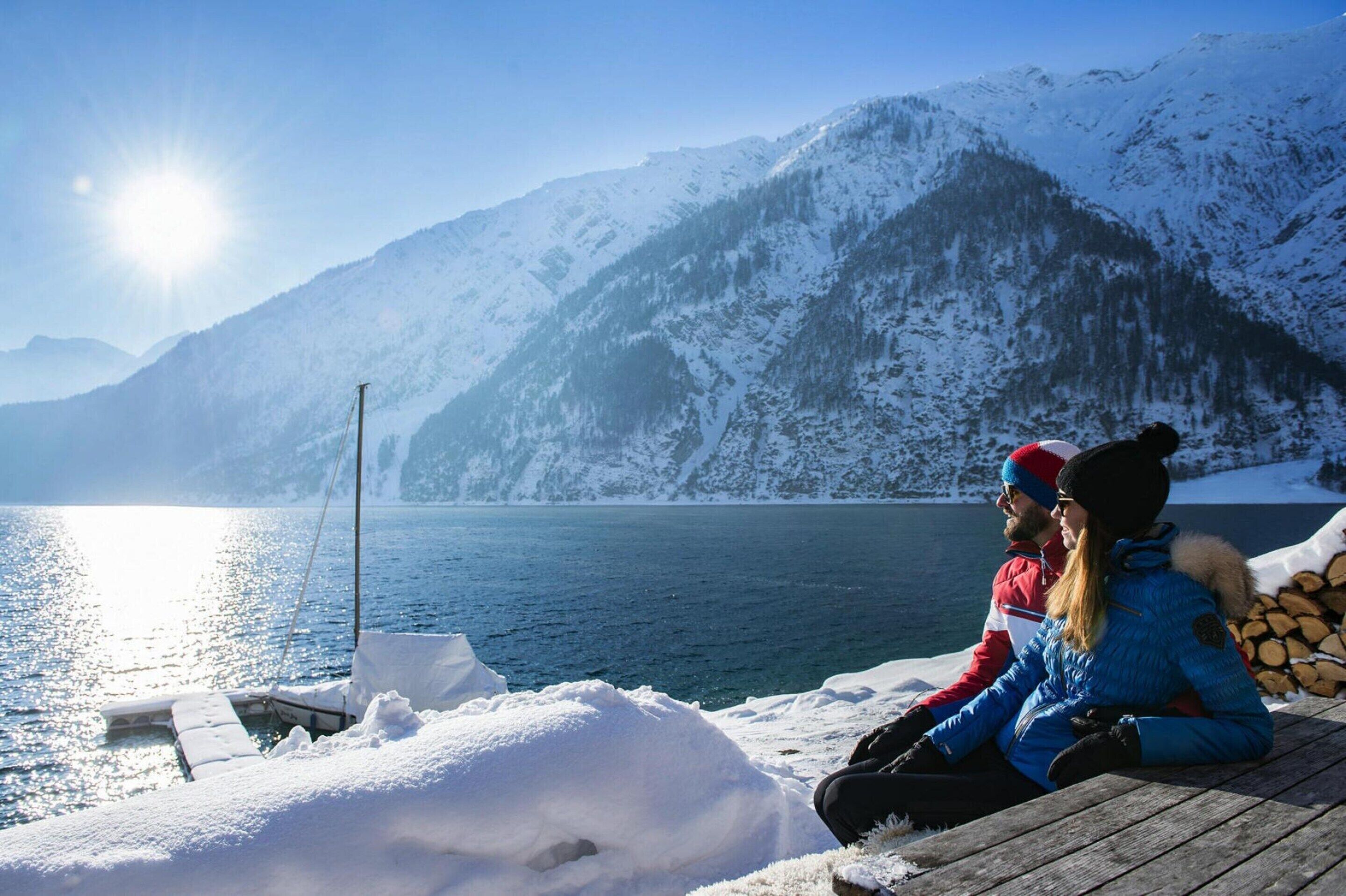 Una pareja sentada en un muelle de madera con vista a un lago tranquilo y montañas nevadas al fondo.