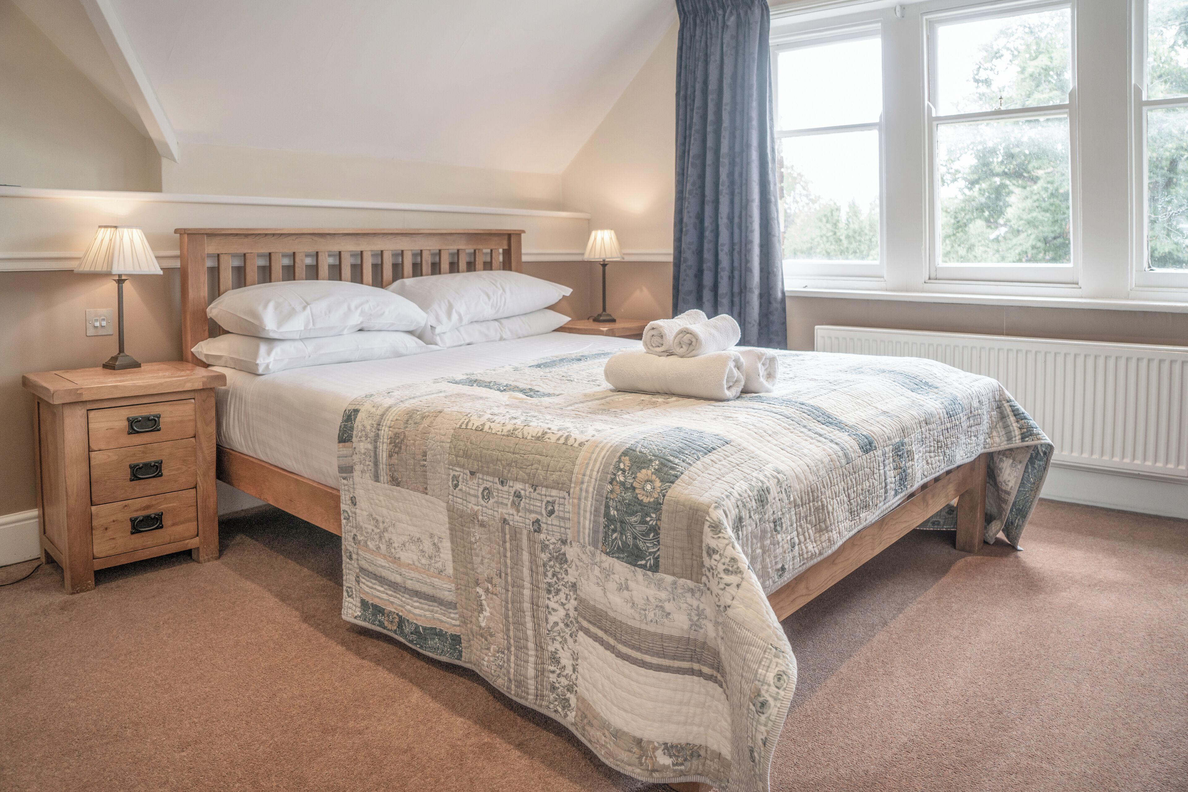 A bedroom with a wooden bed, two bedside tables, a quilt, and a view of trees through a window.