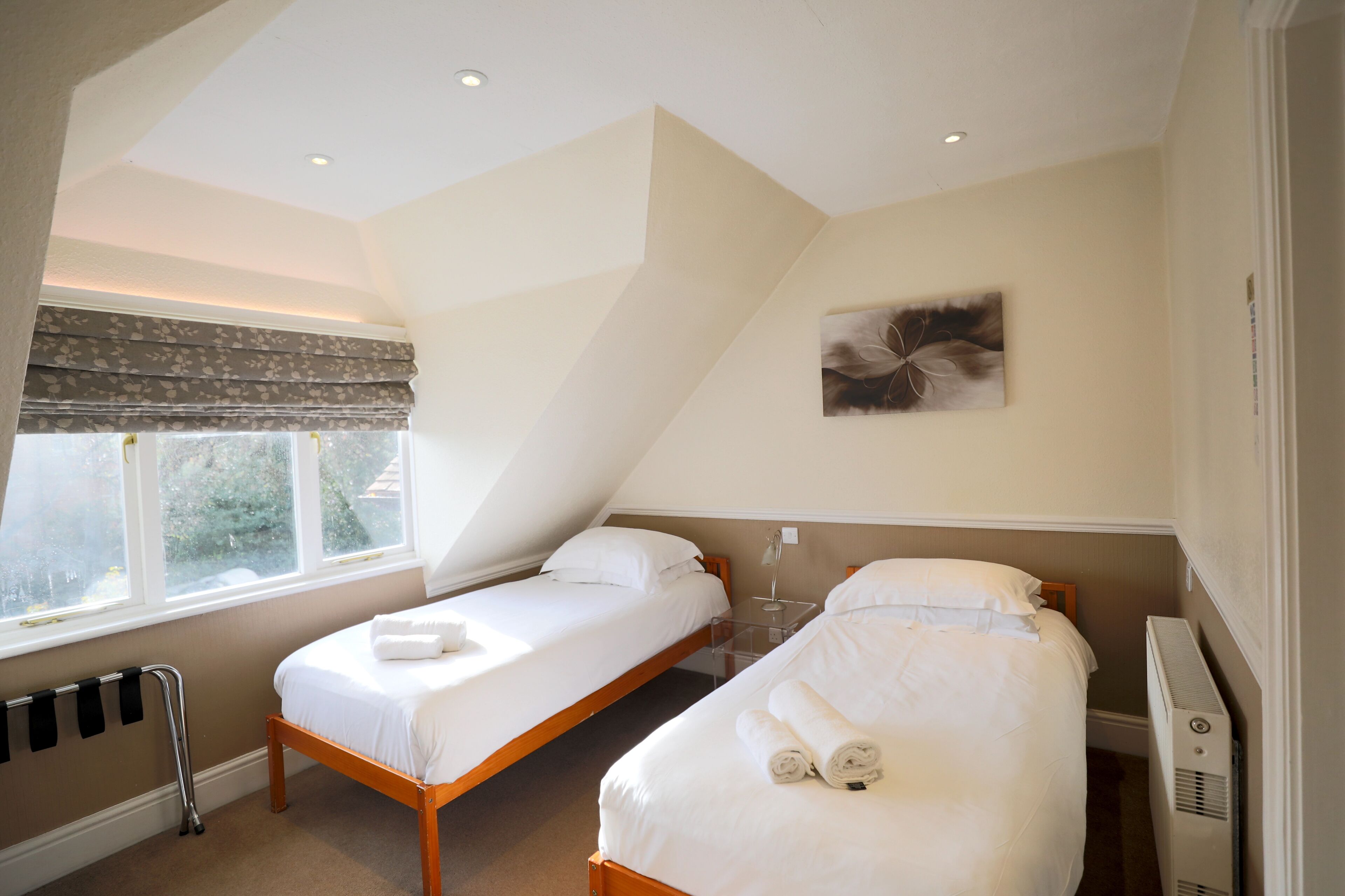 A bedroom with two beds, a radiator, a window with blinds, and a painting on the wall.