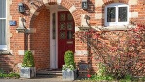 Ein rot backsteiner Gebäude mit roter Tür, einem Erkerfenster mit Buntglas und einem kleinen Garten mit Topfpflanzen.