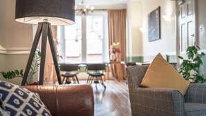 Ein Wohnzimmer mit Stehlampe, einer Couch und einem Sessel. Im Hintergrund ist eine Essecke mit Tisch und Stühlen zu sehen.