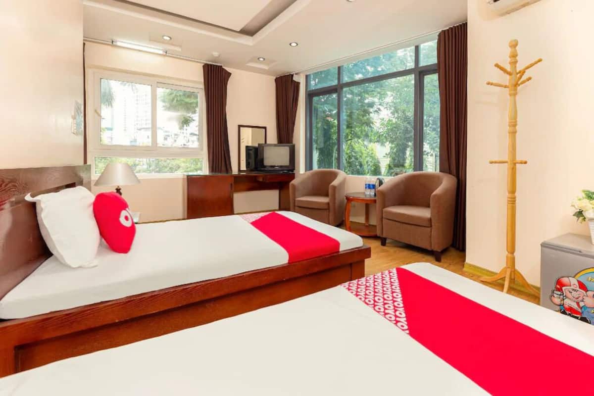 Une chambre d’hôtel comprenant un lit, deux fauteuils, un bureau avec une télévision, un porte-manteau et de grandes fenêtres donnant sur un paysage verdoyant.