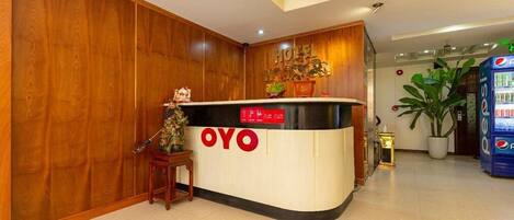 A hotel reception desk with a wooden backdrop, a Pepsi vending machine, and a small plant.