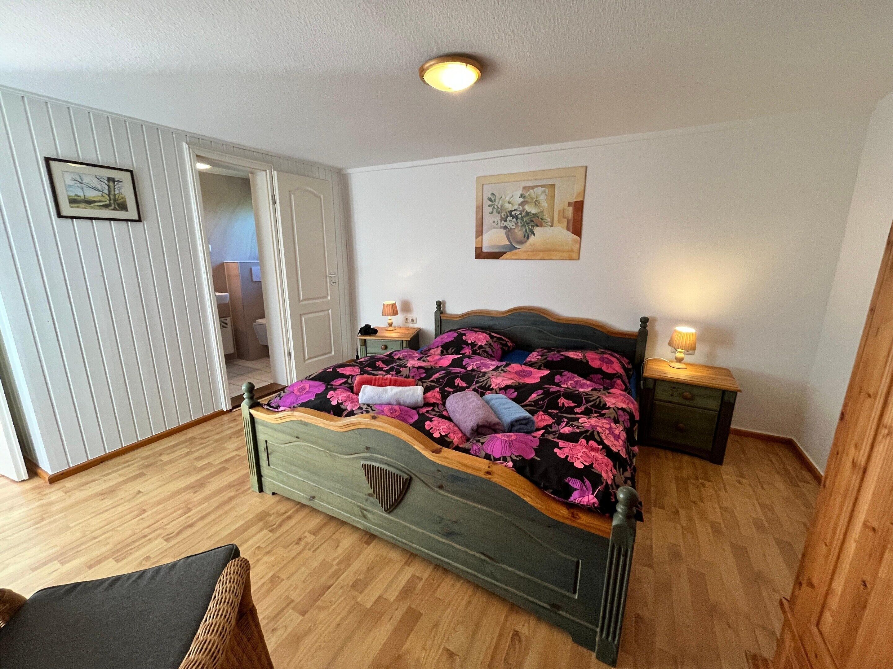 A bedroom with a wooden bed, a nightstand with a lamp, a door leading to another room, and a painting on the wall.