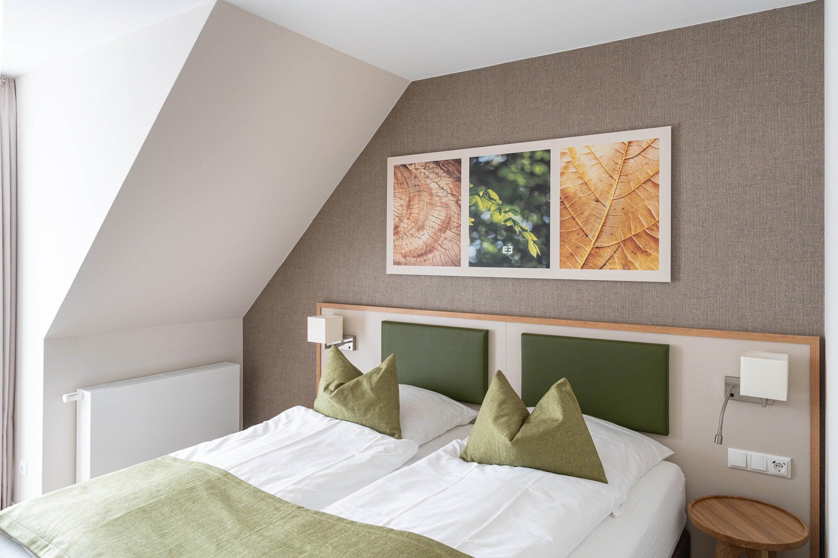 Una habitación de hotel moderna con techo inclinado, cama con ropa de cama verde y blanca, y un cuadro de hojas colgado en la pared.