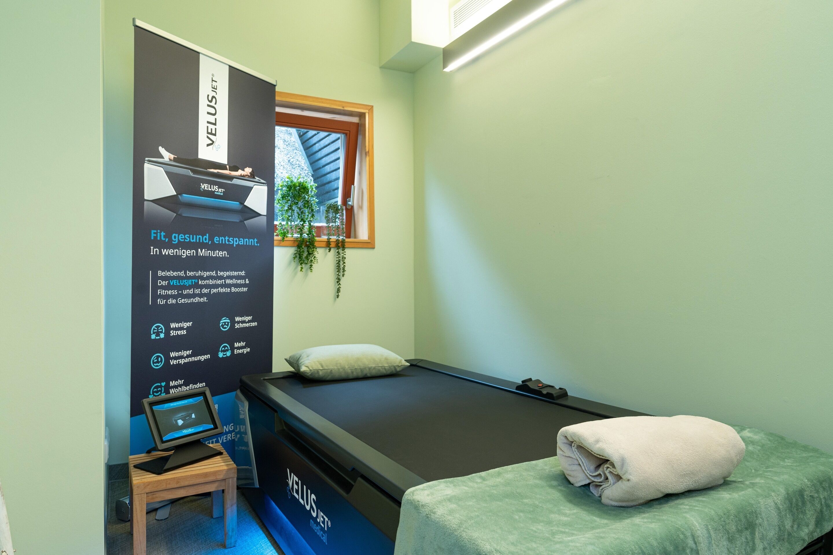 Een massageruimte met een massagetafel, een klein houten krukje met een tablet, en een poster die een welvaartsproduct promoot.
