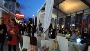 Ein lebhaftes Straßencafé mit Tischen und Stühlen, umgeben von Gebäuden und vielen Menschen.