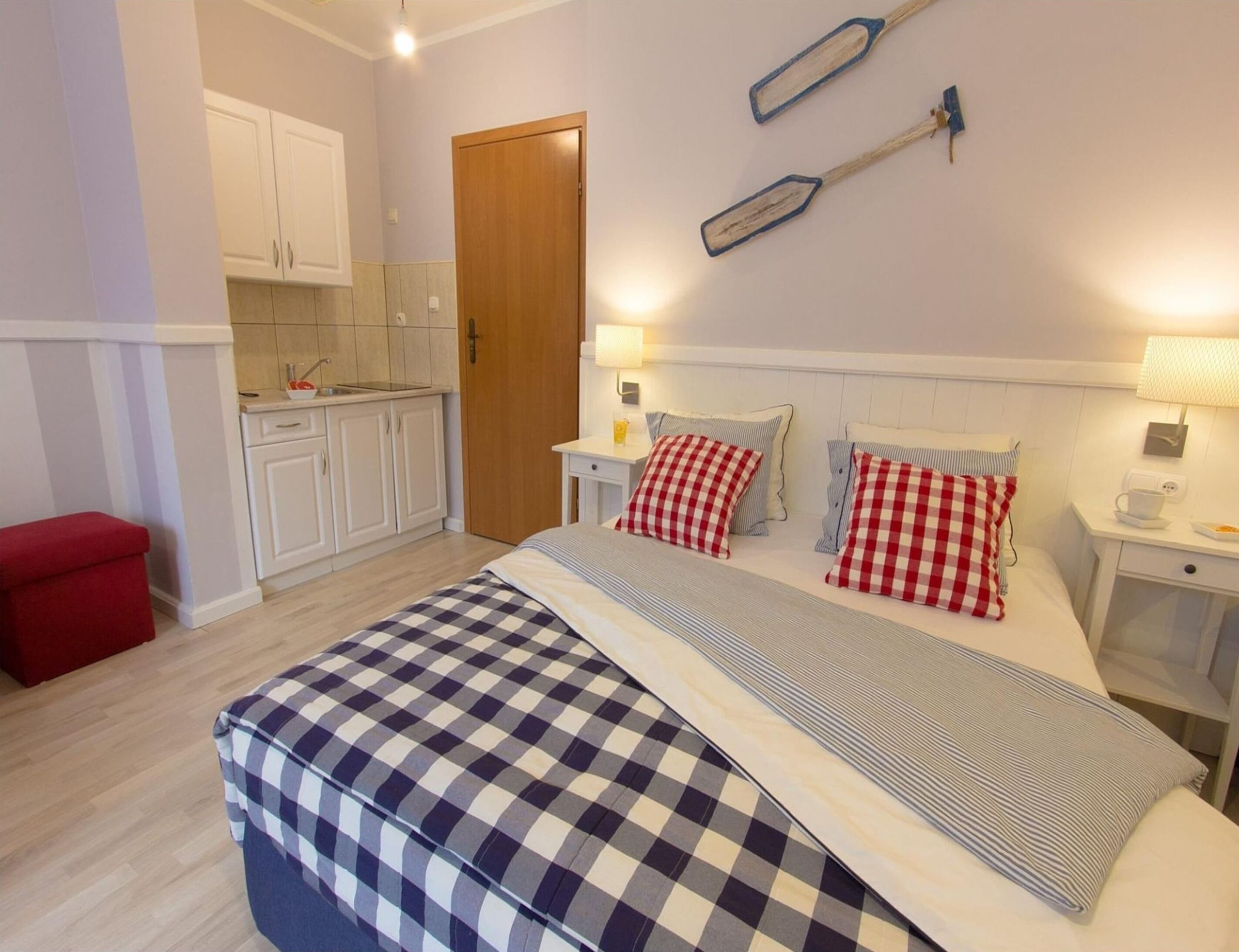 Une chambre à coucher avec une couvre-lit à carreaux, une porte en bois, une petite cuisine avec des placards blancs et un tabouret rouge.