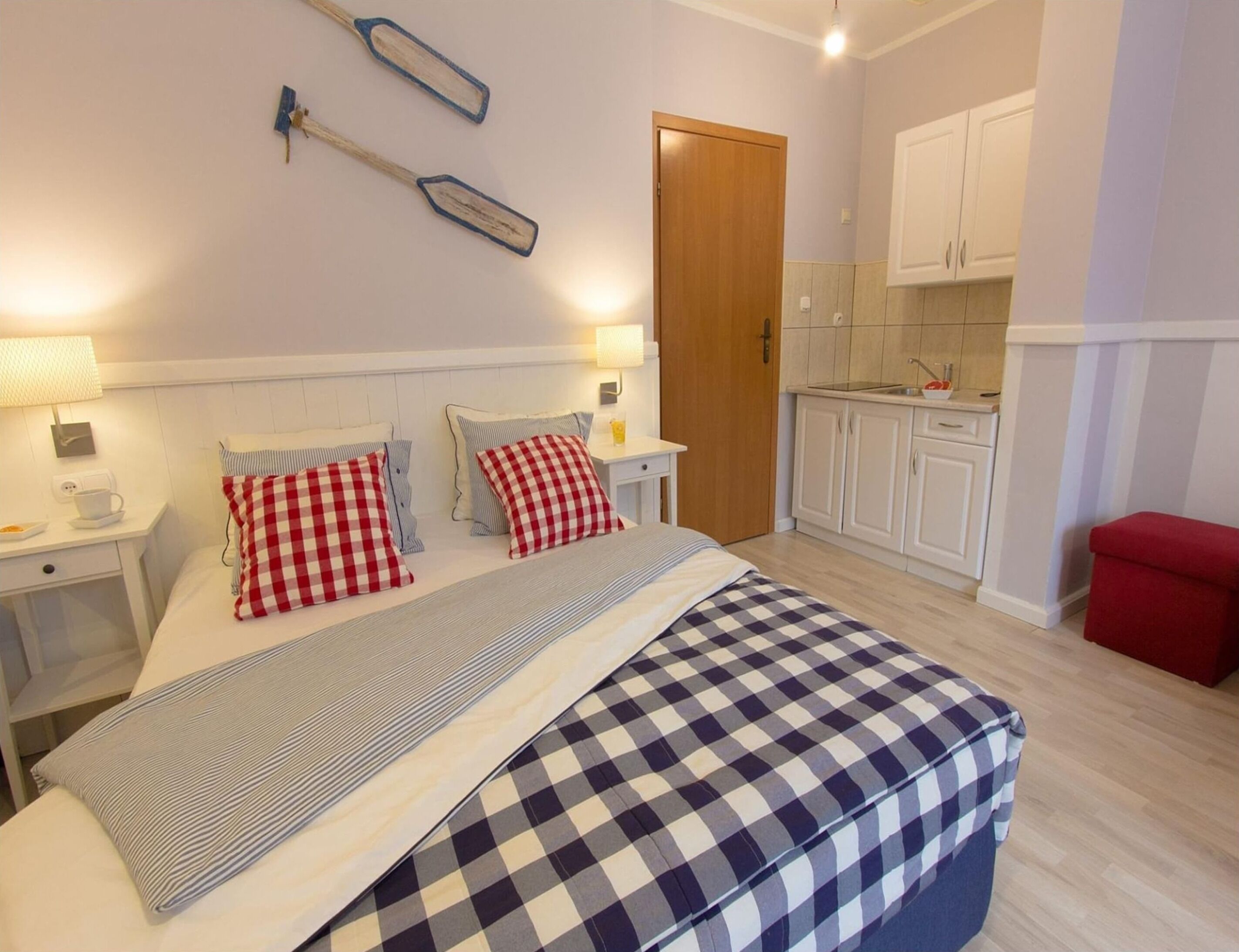 Una camera da letto con un letto, due comodini, una scrivania, una sedia rossa, un angolo cottura con pensili e una porta in legno.