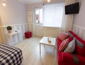 A room with a red sofa, a white coffee table, a red ottoman, a white bedside table, a potted plant, a TV mounted on the wall, and a door with a window. - Apartment for 3 People in Miedzyzdroje (Miedzyzdroje)
