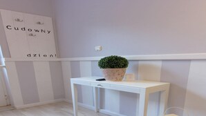 Ein weißes Konsolentische mit einer Topfpflanze und einer Fernbedienung, einer kleinen Laterne sowie einer Wand mit dem Schild 'Cudowny dzient'.