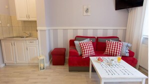 Ein Wohnzimmer mit einem roten Sofa, einem weißen Couchtisch mit Gegenständen darauf, einer Küchenzeile mit Schränken und einem an der Wand montierten Flachbildfernseher.