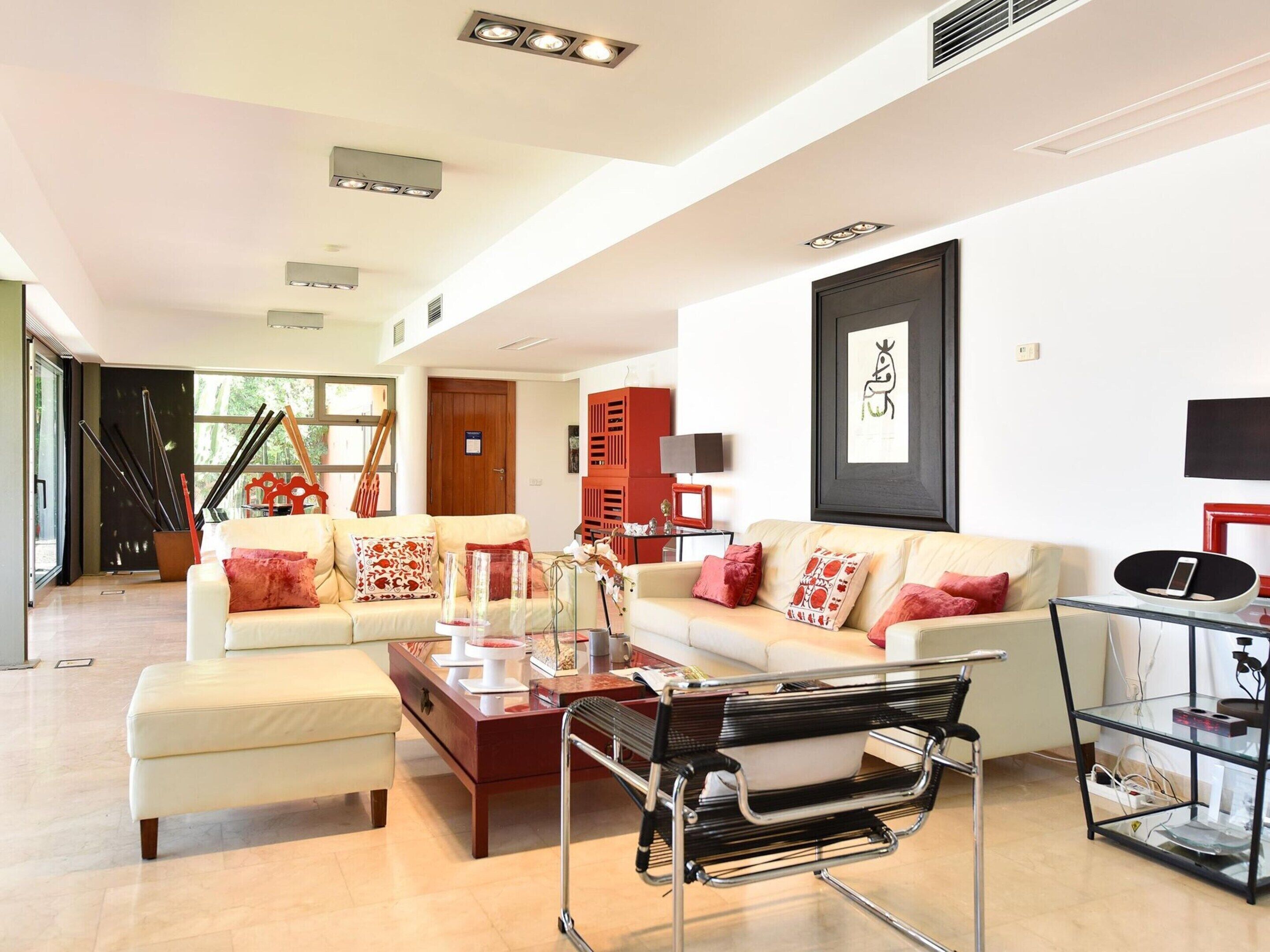 A modern living room with a sofa, ottoman, and a glass coffee table. There is a black metal chair and a glass cabinet with electronic devices.