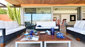 Un espacio al aire libre moderno con mobiliario de mimbre, una mesa de centro de madera con revistas y vistas de la ciudad a través de grandes puertas de cristal.