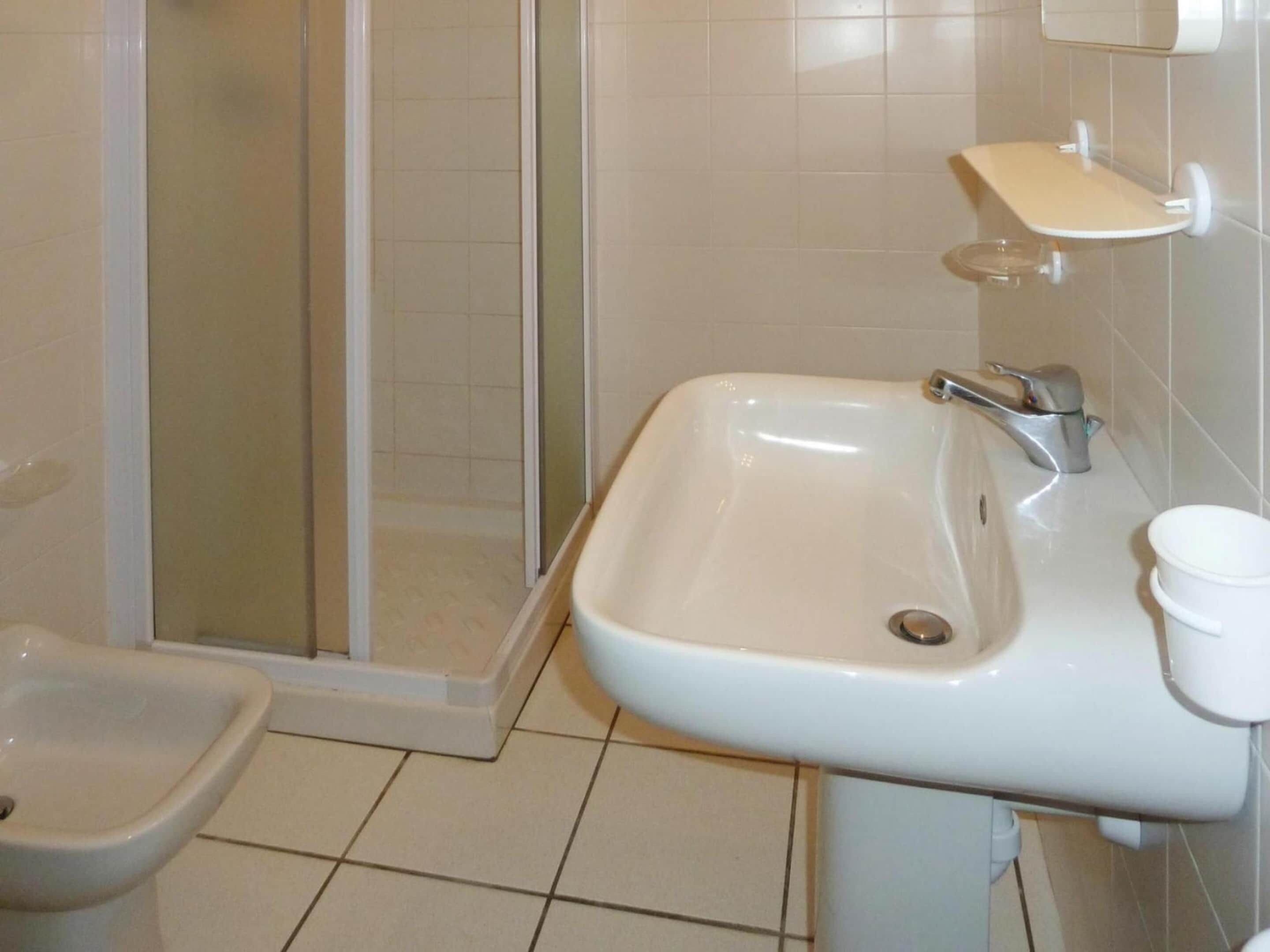 A bathroom with a shower, bidet, sink, and a small shelf.