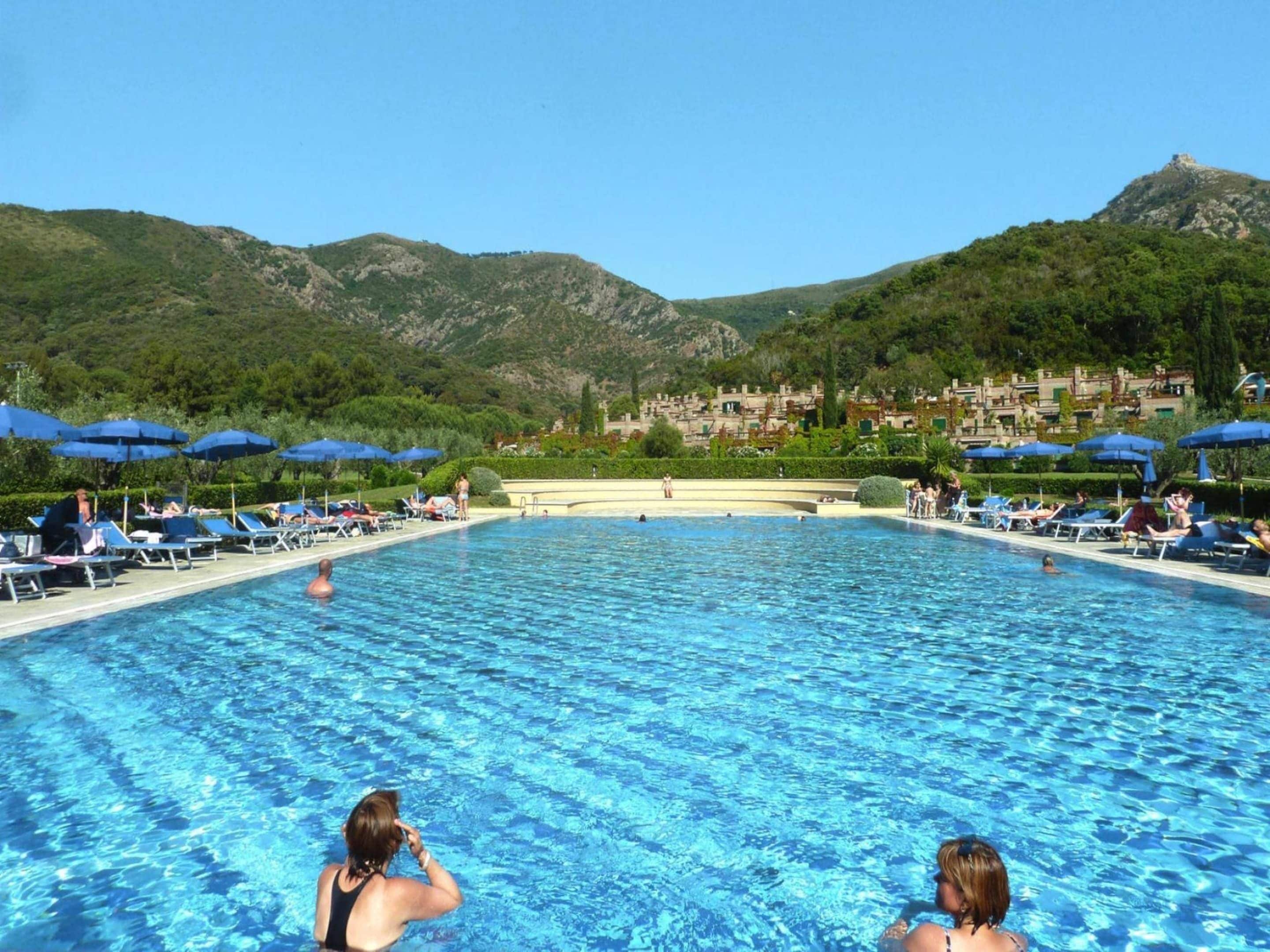 A swimming pool with lounge chairs and umbrellas, surrounded by mountains and a town in the background.