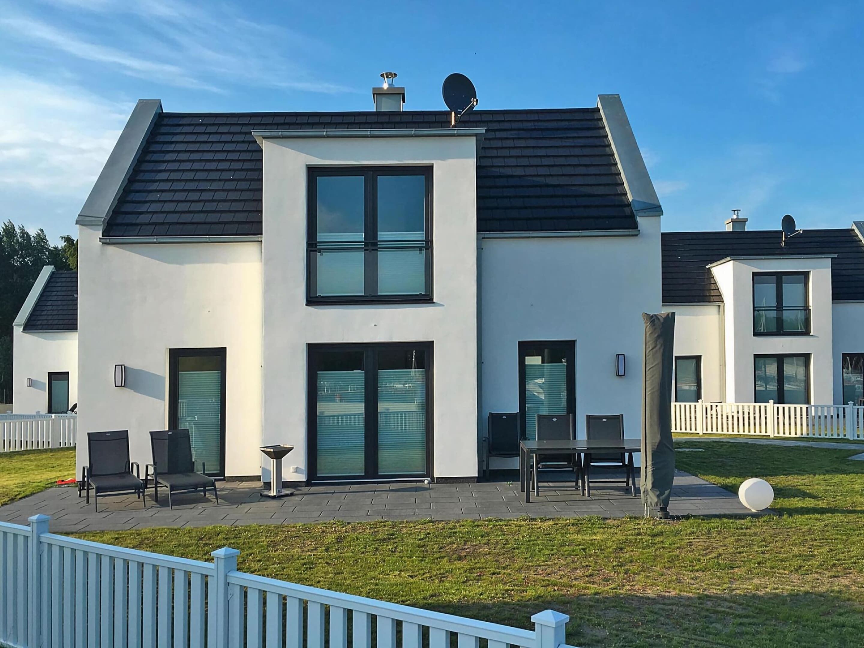 Une maison moderne à deux étages, dotée d’un toit plat, de grandes fenêtres et d’un patio aménagé avec du mobilier d’extérieur.