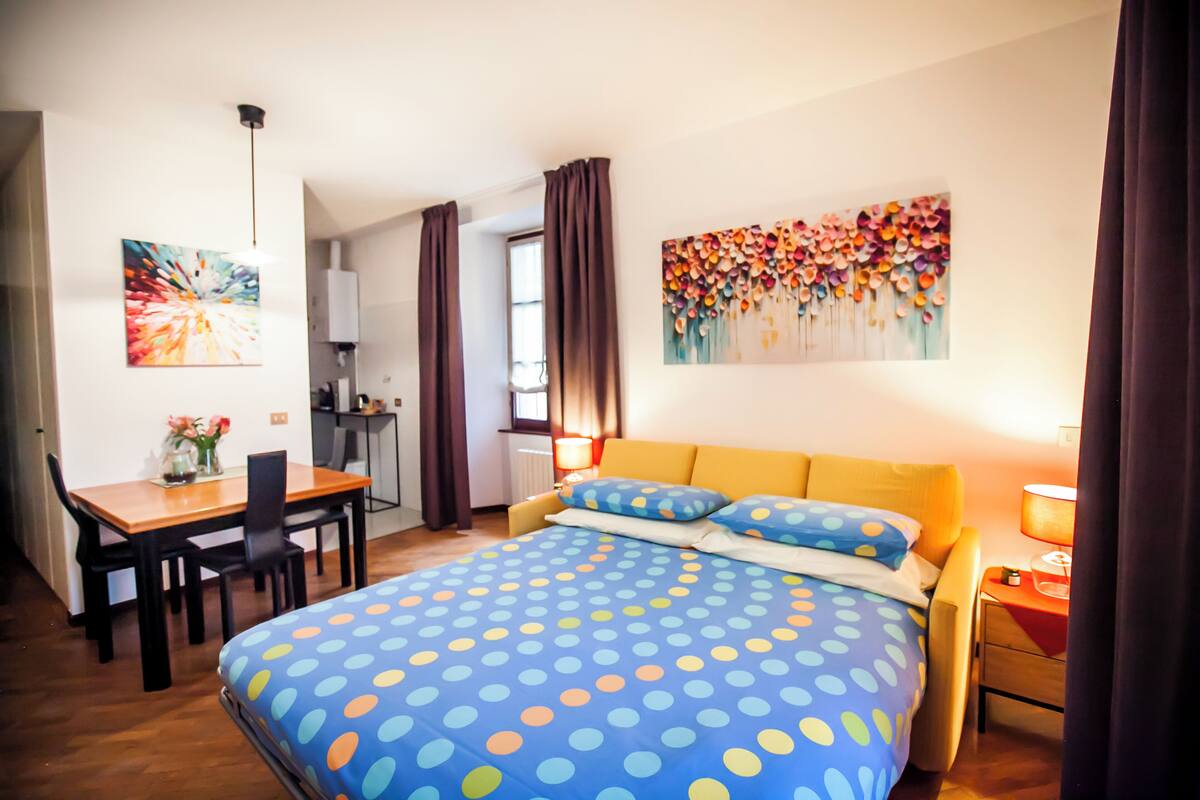Une chambre à coucher avec un lit, un bureau et une chaise. Il y a une fenêtre avec des rideaux, un tableau au mur et une lampe sur la table de chevet.