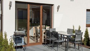 An outdoor patio with a table and chairs, a grill cart, and a sliding glass door leading to an interior space. - Spacious 3-room Apartment, Summer Pool (Postomino)