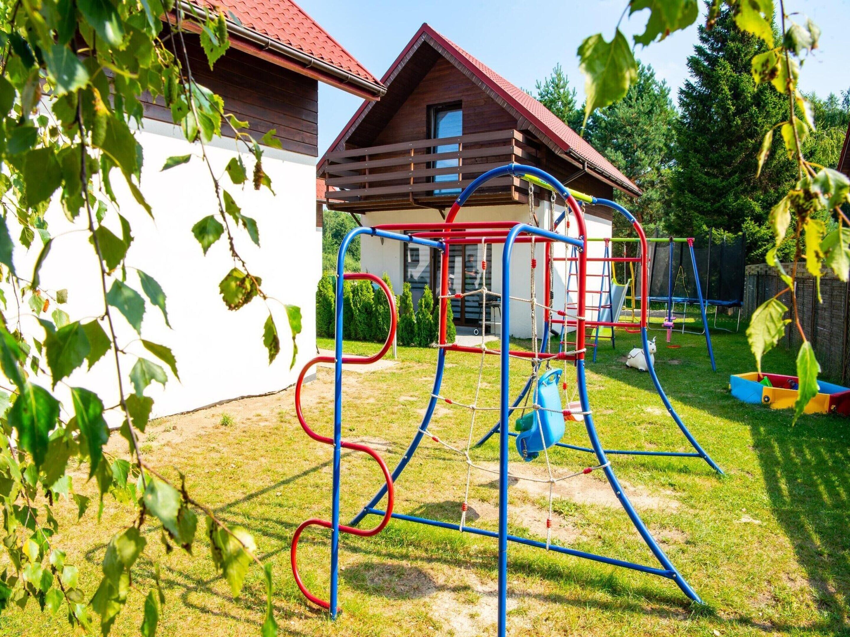 A playground with a swing set and climbing structures, a white building with a red roof, and a grassy yard.