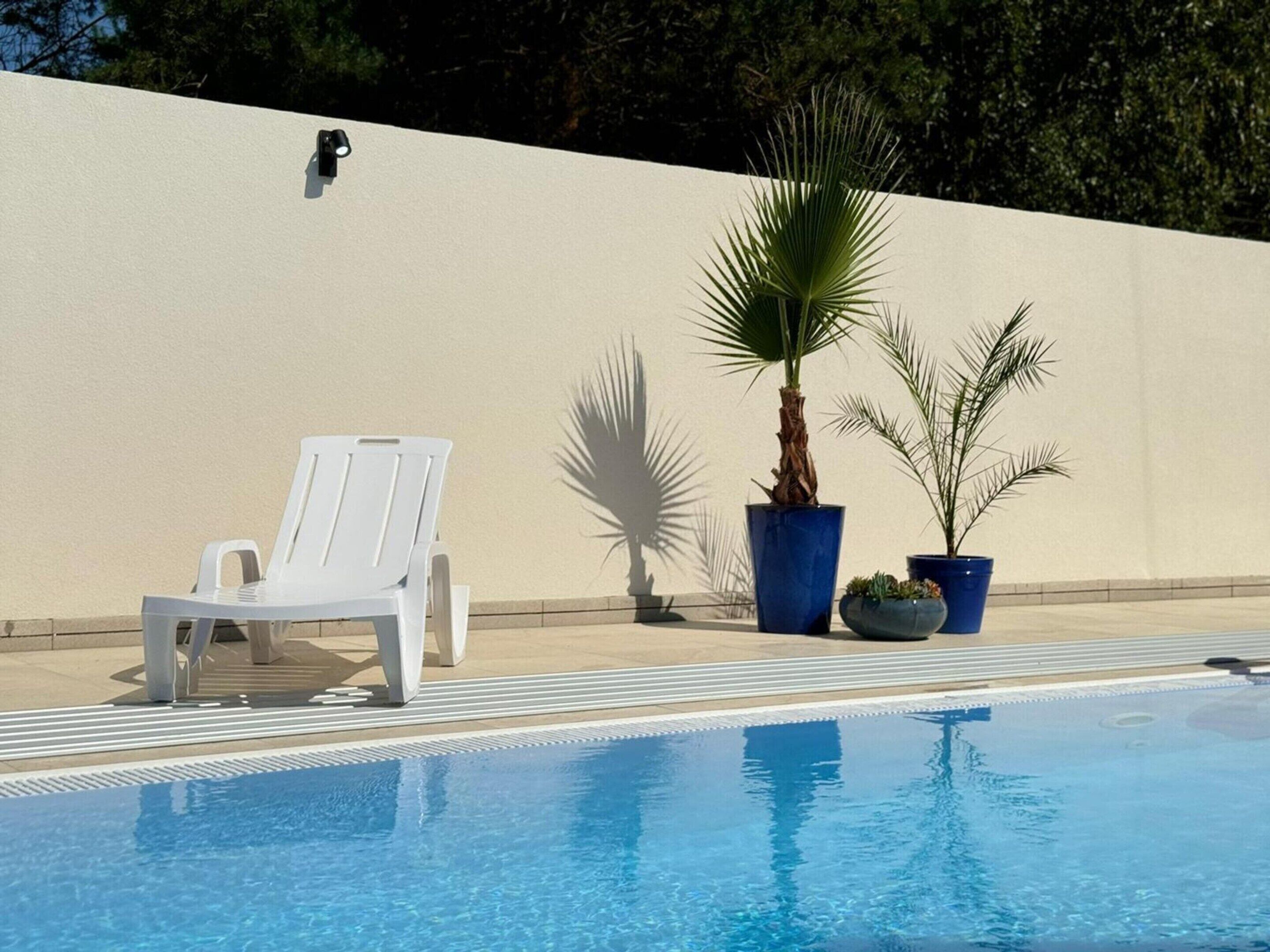 A pool area with a white lounge chair, two potted plants, and a wall-mounted light fixture.