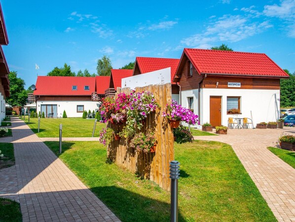 Une rangée de bâtiments aux toits rouges, comprenant un espace d’accueil, un chemin pavé et une clôture en bois ornée de plantes à fleurs.