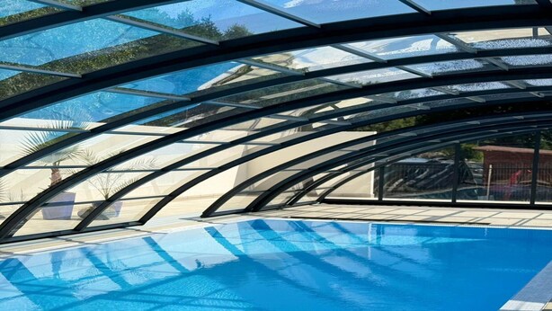A clear view of an indoor swimming pool with a glass ceiling, surrounded by palm trees and a paved deck. - Spacious 3-room Apartment, Summer Pool (Postomino)