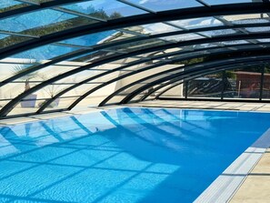 A clear view of an indoor swimming pool with a glass ceiling, surrounded by palm trees and a paved deck. - Spacious 3-room Apartment, Summer Pool (Postomino)