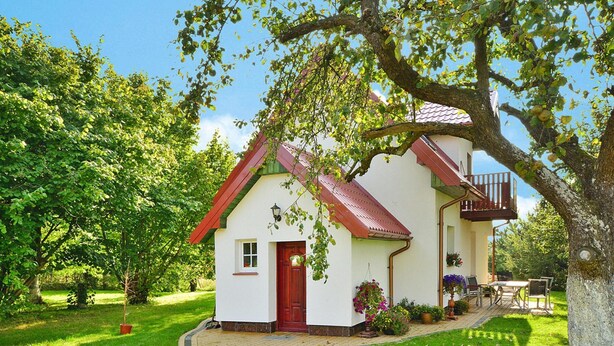 Una casita blanca con puerta roja y un balcón, rodeada de vegetación exuberante y un sendero empedrado.