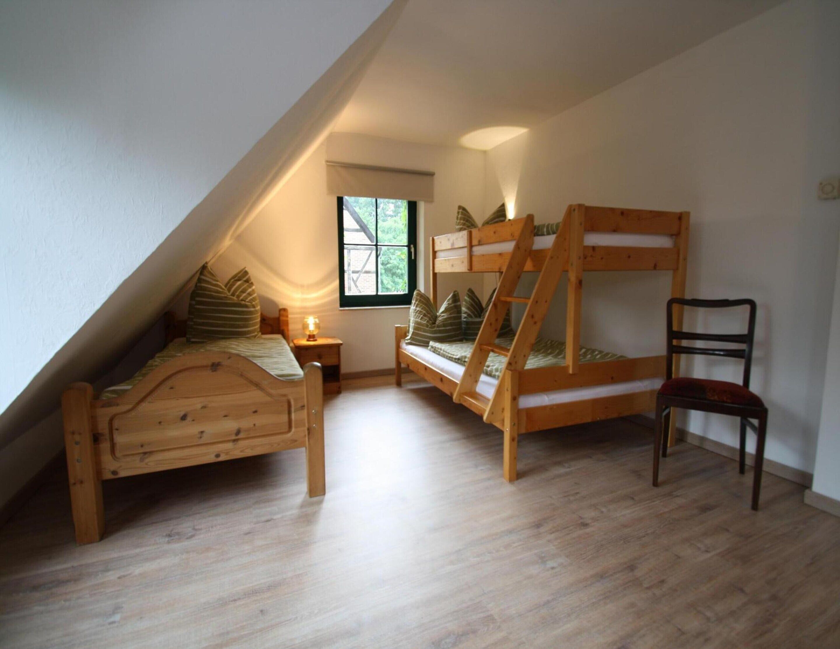 A cozy attic bedroom with a sloped ceiling, a wooden bunk bed, a single bed, a chair, and a small table with a lamp.