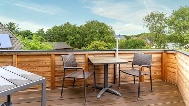 Eine Terrasse im Freien mit Tisch und zwei Stühlen, umgeben von Holzwänden und Blick auf Bäume und Häuser.