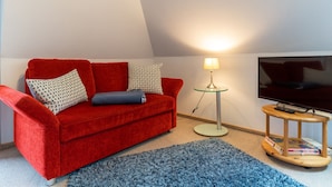 Ein gemütliches Wohnzimmer mit einem roten Sofa, einem Glastisch mit einer Lampe, einem Fernseher und einem kleinen Holztisch mit Büchern.