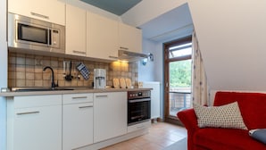Eine kleine Küche mit Mikrowelle, Backofen und Kaffeemaschine. Ein rotes Sofa mit einem gemusterten Kissen ist zu sehen.