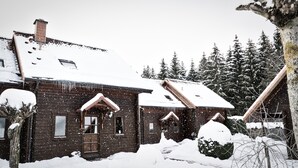 Una cabaña de madera con techo nevado y una chimenea, rodeada de nieve y árboles.