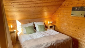Un dormitorio acogedor tipo cabaña de madera con techo inclinado, una cama con colcha color beige, dos lámparas de noche y un cuadro enmarcado en la pared.