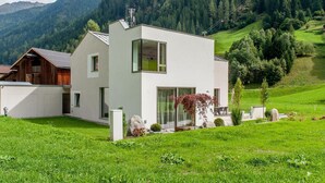 Una casa moderna de dos pisos con un amplio ventanal, rodeada por un exuberante paisaje verde y montañas al fondo.