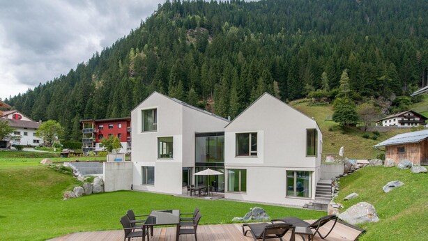 Una casa moderna de dos pisos con un balcón, rodeada por un paisaje exuberante y un telón de fondo montañoso boscoso.
