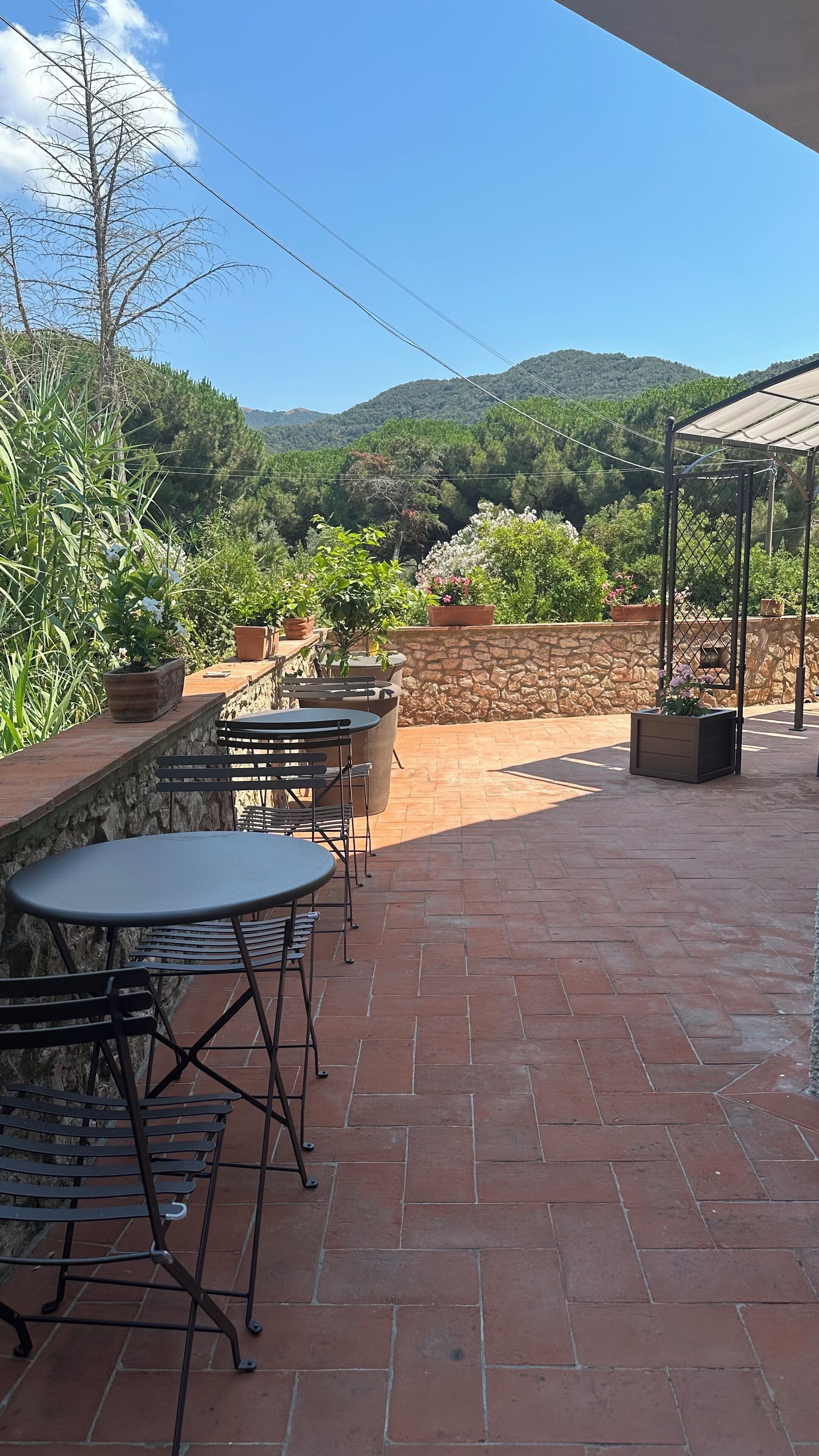 An outdoor patio with terracotta-tiled flooring, metal tables and chairs, potted plants, and a stone wall.