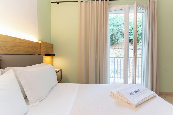 A hotel room with a bed, a nightstand, a lamp, and a view of the outdoors through a window with curtains. - Elbitat Homes (Rio)