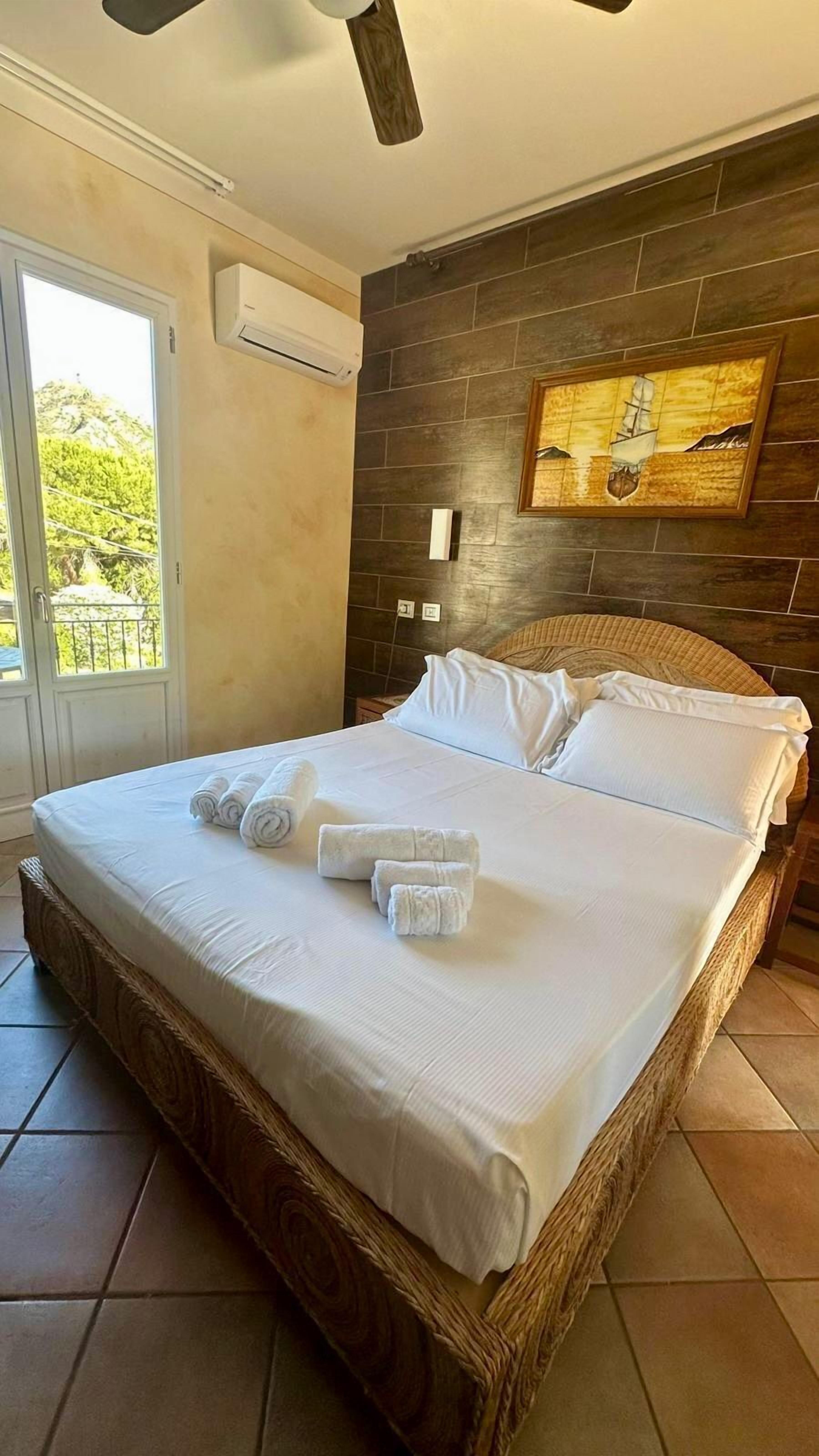 Una cama bien hecha con ropa de cama blanca y toallas enrolladas, cabecera de madera, ventana grande con vistas a la vegetación y un aire acondicionado instalado en la pared.