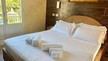 Una cama bien hecha con ropa de cama blanca y toallas enrolladas, cabecera de madera, ventana grande con vistas a la vegetación y un aire acondicionado instalado en la pared.