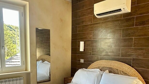 A bedroom with a bed, a ceiling fan, an air conditioning unit, a window, and a wall-mounted mirror. - Elbitat Homes (Rio)