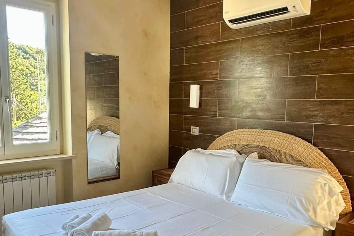 A bedroom with a bed, a ceiling fan, an air conditioning unit, a window, and a wall-mounted mirror.