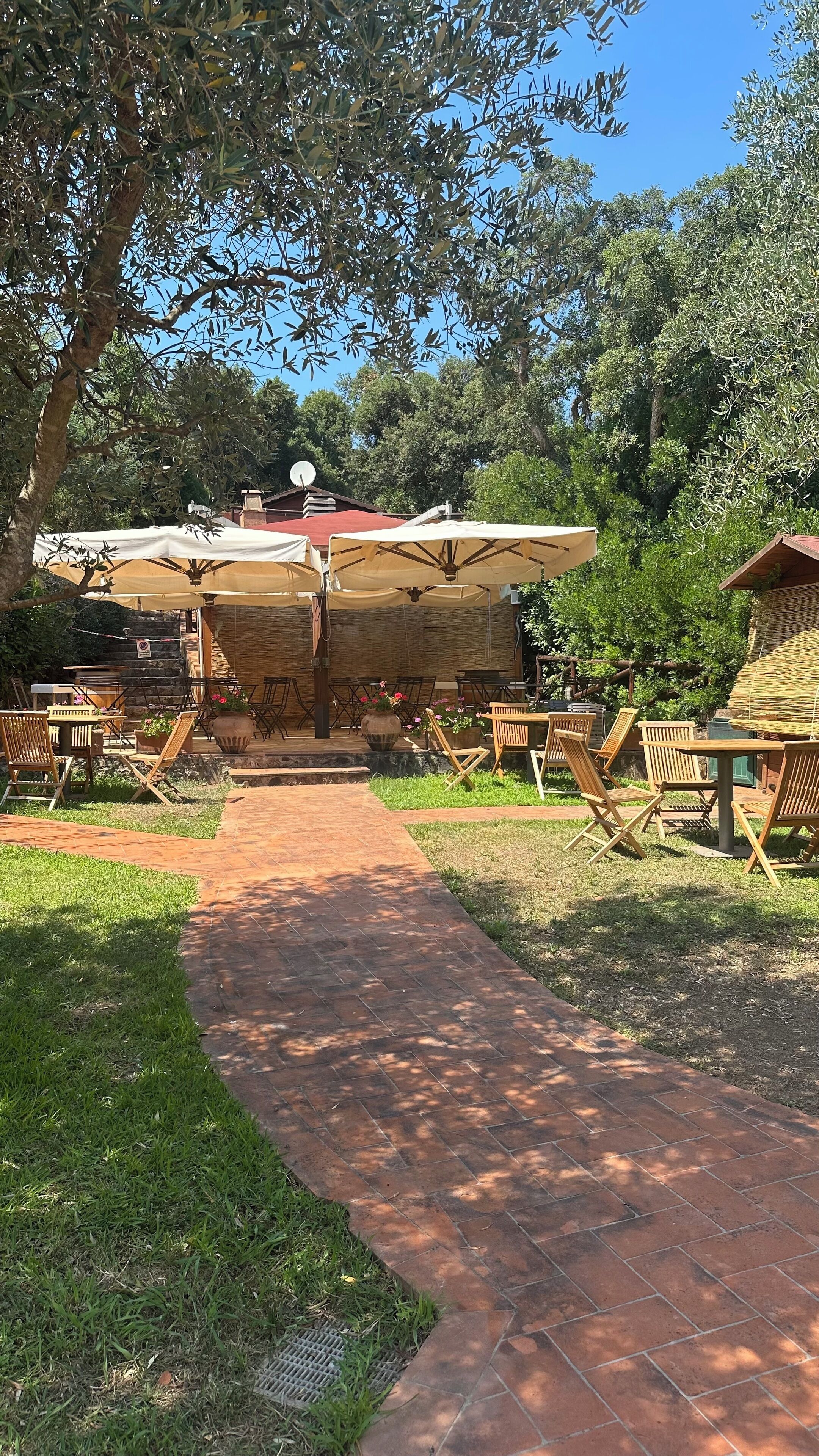 Un patio al aire libre con suelo de ladrillo, mesas y sillas de madera, y un área de estar cubierta con sombrillas.